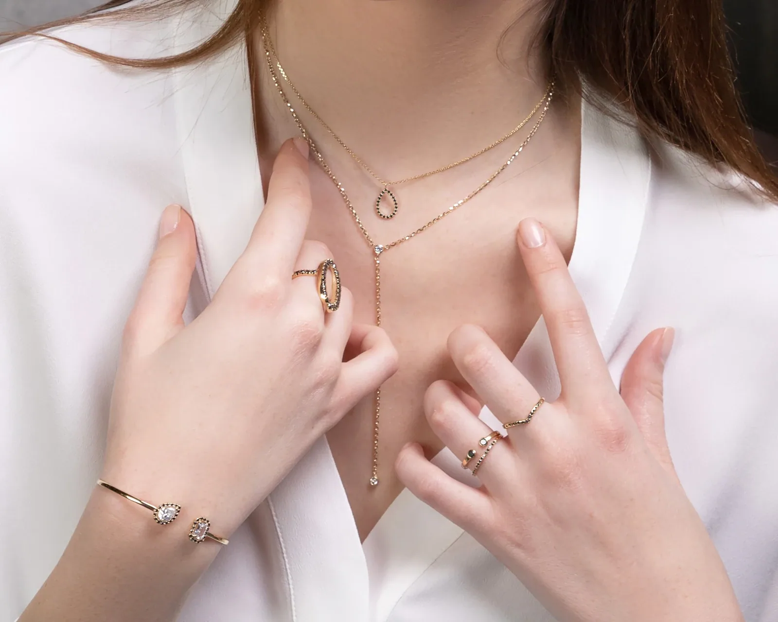 Woman in a white blouse wearing layered gold necklaces, stacked rings and an open cuff bracelet